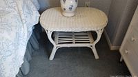 Side view of the white painted wicker coffee table placed beside a bed and near a white drawer. Shows detail of weaving and structure.