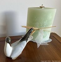 White ceramic bird figurine with gray and black detailed wings and head, positioned next to a large green oil candle tied with bow, sitting on wooden surface