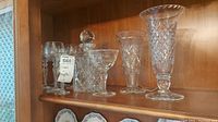Seven wine glasses, crystal decanter with stopper, two glass vases, and crystal goblet arranged on a wooden shelf.
