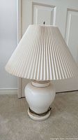 Full view of the vintage ceramic lamp with white pleated fabric shade standing on carpet in front of a door.