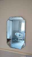 Front view of the rectangular bevelled mirror reflecting a wicker chair and part of a window with curtain.