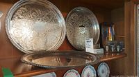 Three ornate round silver plated trays on a shelf with additional items behind