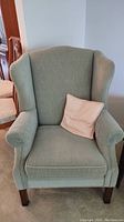 Full view of the light green upholstered wingback chair with a peach-colored pillow on the seat.