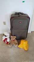 Gray Samboro suitcase standing upright with two plush toys and yellow hat in front on carpet