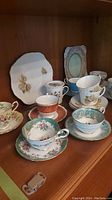 Photo showing ten tea cups and saucers and the cake plate arranged on a wooden shelf.