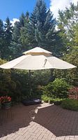 Outdoor view of large beige patio umbrella fully opened on a brick patio with green trees and plants in background.