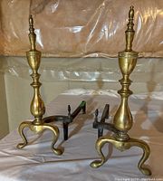 Pair of vintage brass and iron andirons showing front and left sides, set on cloth background in a basement setting with plastic sheeting on walls.