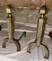 Pair of vintage brass and iron andirons with hammered texture and curved iron legs on a concrete floor.