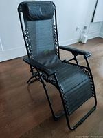 Zero gravity recliner chair positioned indoors on a wood floor, showing mesh fabric seating surface, black metal frame, lacing attachment, and padded armrests with headrest pillow.