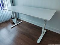 White IKEA adjustable desk showing the left angled side with metal legs and a crank wheel for height adjustment, placed on a wooden floor near a window.