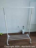 Front view photo of a white metal clothes rack showing the hanging bar and lower slatted shelf on wheels.