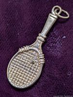 Close-up photo of the gold tennis racket pendant showing detailed grid pattern strings and tennis ball attached to the racket face.