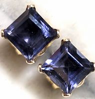 Close-up front view of two square-cut iolite gemstone stud earrings showing deep bluish-purple stones with gold prong settings.