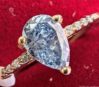 Close-up of pear shaped fancy blue lab grown diamond set in three prongs on yellow gold band with round accent stones