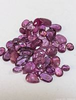 Close-up photo of approximately 39 loose genuine rubies in various shapes and sizes, with purple to pinkish-red tones on white background.