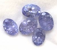 Close-up of five oval faceted tanzanite gemstones arranged loosely on a white surface showing their light to medium violet-blue color with internal inclusions.