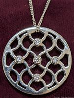 Close-up photo of the round silver pendant showing the lattice design and embedded cubic zirconia stones on the chain.