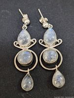 Front view of the pair of sterling silver drop earrings set with pear-shaped and round moonstone cabochons against a black background.