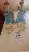 Front view of two vintage Fairylite lady figurines with matching blue dresses and pink hats, each accompanied by a small white leaded poodle dog figurine.