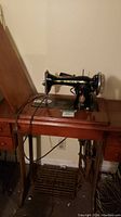 Antique Singer sewing machine mounted in a wooden cabinet table with drawers and pedal mechanism. Photo shows machine side profile and cabinet top.