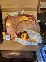 Three copper serving pieces displayed in a cardboard box. The large rectangular tray features intricate etched and hammered patterns. The round platter shows a hammered texture. The smallest tray has a scalloped edge and both smaller pieces are partially wrapped in white protective cloth.