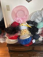 Photo showing a stack of assorted women's hats in various colors including pink, black, red, blue, yellow, and beige. Some have bows or floral decorations. Hats mostly formal style and appear gently used or new.
