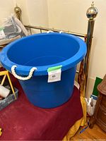 Large blue plastic tub container with rope handles sitting on a red cloth surface near brass bedposts.