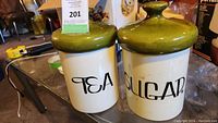 Pair of ceramic canisters labeled TEA and SUGAR with green mushroom-shaped lids on a table.