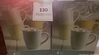 Photo of two sealed boxes, each containing 4 mugs. One box has light gray mugs, the other pastel yellow. Both boxes show 4 mugs with handles and a simple design.