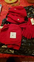 Three red fleece hats with logo patches, red fleece gloves, and red fleece scarf displayed on black wrapping with original tags attached.