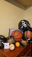 Photo showing two black and white skull design helmets, a boxed Rawlings basketball, Franklin adult size football, two black baseball gloves with baseballs inside, one yellow softball, and one loose baseball on a wood surface.
