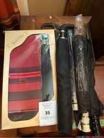 Photo shows three umbrellas on a wooden surface: one in original packaging with red and black stripes, one black umbrella with curved handle, and one black umbrella still in plastic wrapping.
