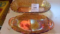 Two oval amber carnival glass dishes shown on table; one smooth with etched diamond pattern, one with handles and embossed floral pattern.