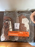Two vintage moccasin making kits in clear plastic packaging with brown suede and laces visible, one labeled 'Ladies Large'.