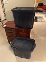 Two Rubbermaid Roughneck plastic storage bins with lids stacked and placed on wooden furniture, showing their size and color.