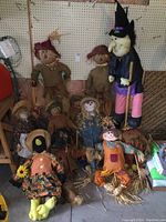 Wide group photo showing all scarecrows and large witch figure together grouped against a wall.