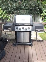 Front view of Broil King stainless steel propane BBQ grill showing control panel with four knobs, grill lid with temperature gauge, side shelves, and wheels.