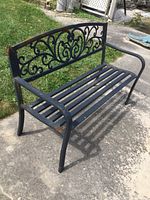 Front-side angle view of black metal bench with ornate backrest on concrete surface near grass.