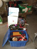 Blue plastic bin filled with assorted Christmas decorations including wreaths, Santa figures, ornaments, mini sleigh, and storage totes visible behind.