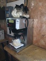 Front and side views of a commercial grade stainless steel coffee maker with glass pot filled with paper filters and two additional glass pots on top also containing paper filters.