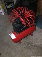 Small red portable air compressor with cylindrical tank and black motor housing, featuring coiled red air hose attached on top.