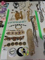 Top view of assorted costume jewelry items including necklaces, bracelets, brooches, rings, cuff links, and belt buckles arranged on a white surface with green tape lines.