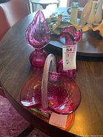 Photo showing three cranberry glass pieces on a table including a basket with handle, a vase, and a bird-shaped piece.