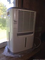 Front-side view of white Frigidaire dehumidifier positioned by a window, showing air vents and water level indicator.