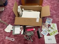 Nintendo Wii console boxed setup with sensor bar, remotes, cables, game discs, and manuals laid out on carpet.