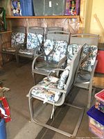 Six metal patio chairs with floral cushions and three matching metal side tables stored in a garage.