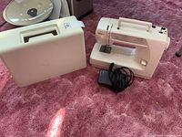 Full view of sewing machine next to white carrying case and black foot pedal on pink carpet