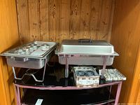 Two metal steam servers on a black countertop against a wooden wall, one with lid on, one without. Boxes with sterno cans and holders next to them.