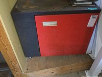 Closed metal storage cabinet with red door and black body on a wooden platform.