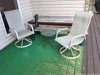 Two light beige patio swivel chairs with metal frames and a small round metal side table on an artificial green grass mat beside a wooden bench and patio deck.